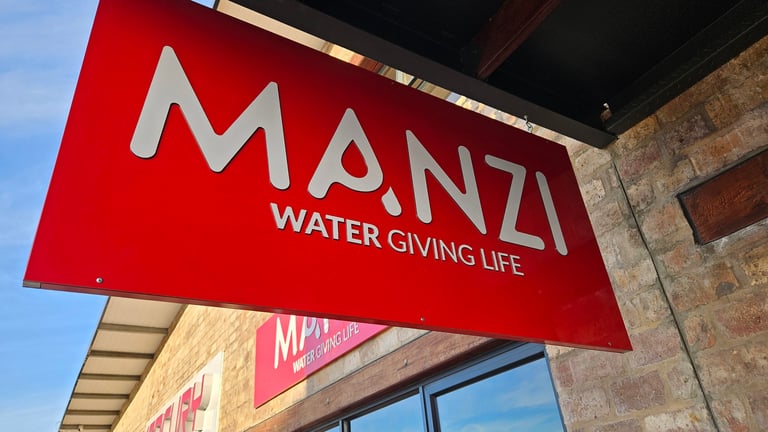 Red sign with white MANZI logo and WATER GIVING LIFE text hanging outside a building storefront